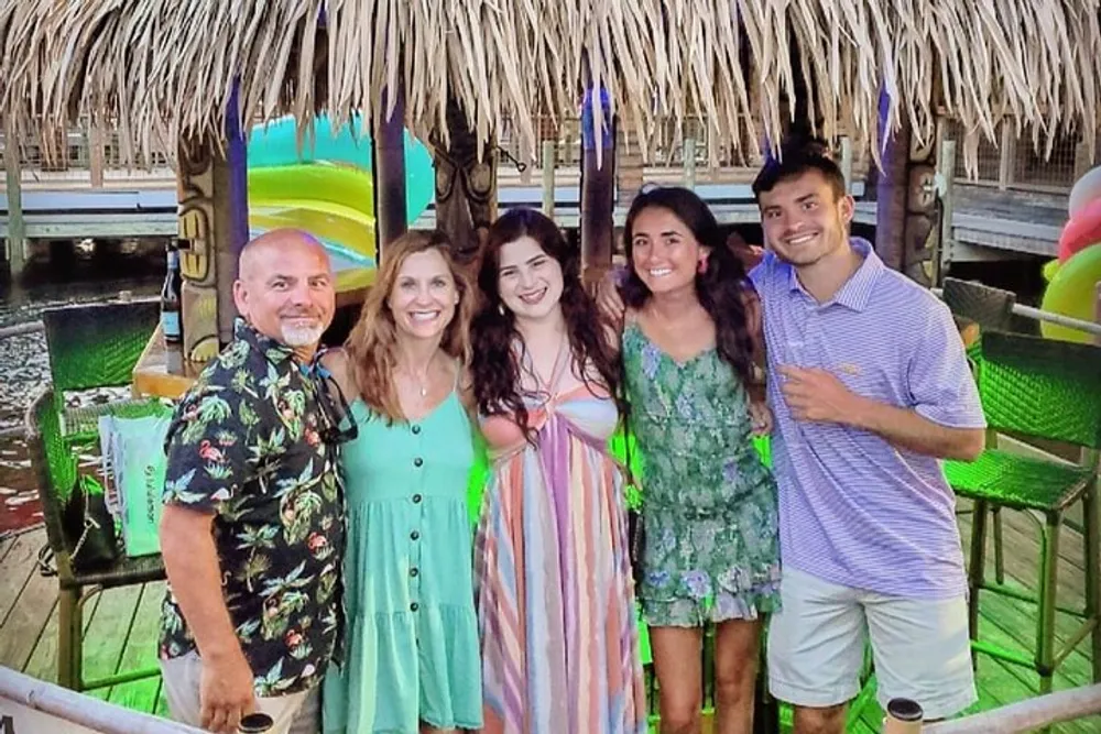 Five people are smiling for a group photo at a tropical-themed outdoor venue with tiki-style decorations and a water slide visible in the background