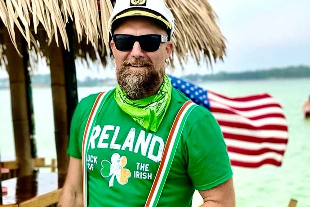 A person is wearing a green Ireland t-shirt with a captains hat sunglasses and a green bandana standing in a tropical setting with an American flag in the background