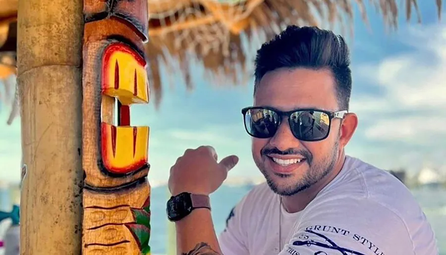 A smiling person is pointing at a colorful wooden tiki sculpture while wearing sunglasses and a wristwatch, with a beach setting in the background.