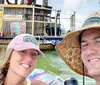 A man and a woman wearing hats are smiling for a selfie with a colorful tube and a boat in the background suggesting they are enjoying a day at a waterfront or marina