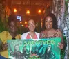 Three people are smiling and holding a sign stating WE SURVIVED with a horror-themed backdrop that includes eerie green lighting and skulls suggesting they completed a scary entertainment attraction