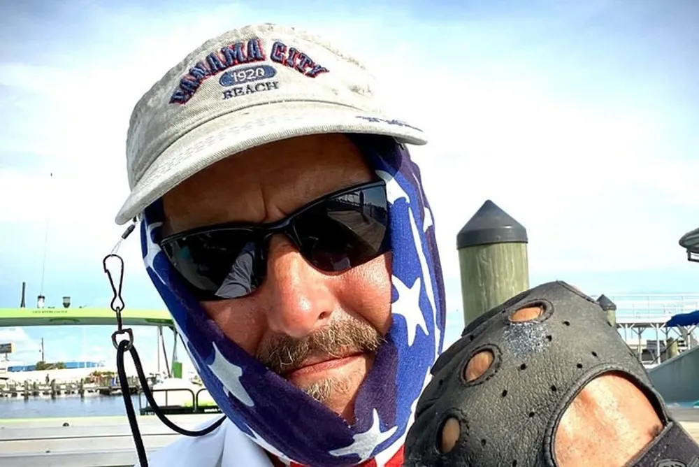 The image shows a person sporting sunglasses a cap and a star-spangled bandana with a fishing hook attached near the brim of the cap posing for a selfie outdoors