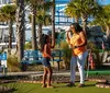 A family of four is enjoying a game of mini-golf on a sunny day with a ferris wheel and other attractions in the background