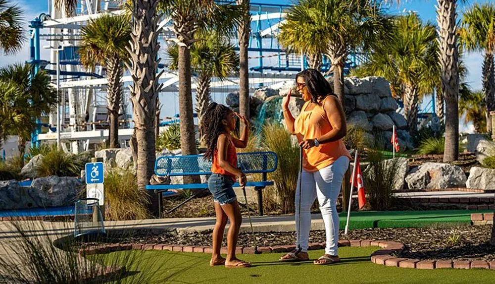 Two people are enjoying a sunny day playing miniature golf with amusement park rides in the background