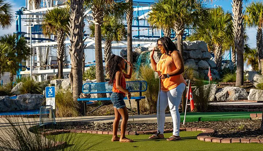 Two people are enjoying a sunny day playing miniature golf with amusement park rides in the background.