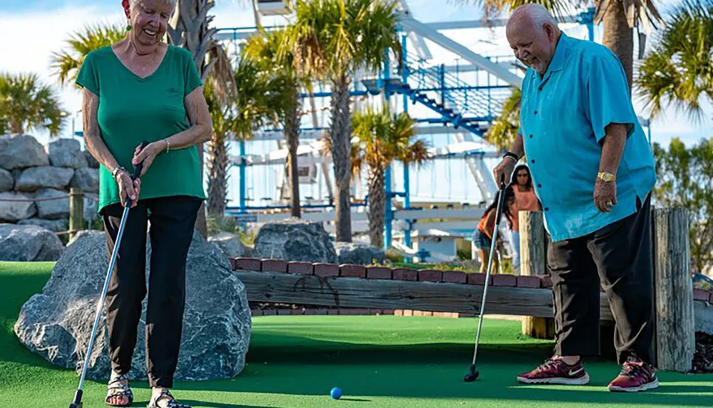 Two older adults are enjoying a game of mini-golf on a sunny day with palm trees and a roller coaster in the background