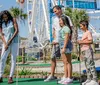 A family of four is enjoying a game of mini-golf on a sunny day with a ferris wheel and other attractions in the background