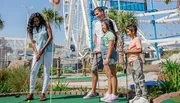 A family of four is enjoying a game of mini-golf on a sunny day with a ferris wheel and other attractions in the background.