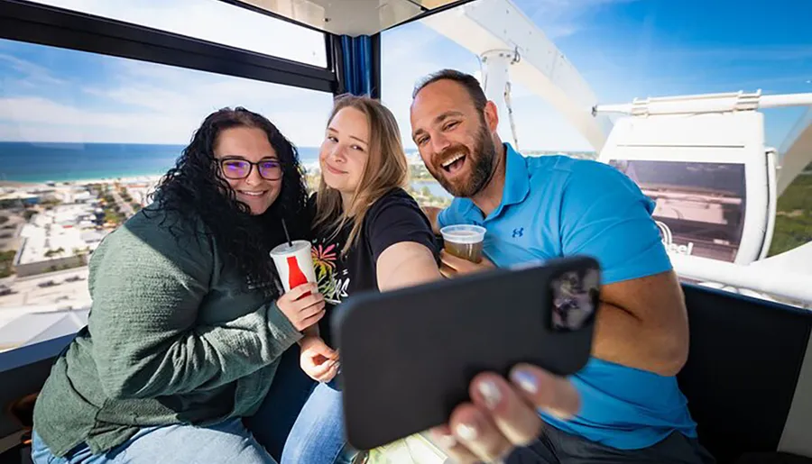 Three people are taking a selfie together while riding on a Ferris wheel, displaying a scenic view of the coastline in the background.