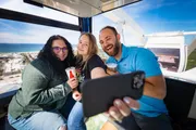 Three people are taking a smiling selfie inside a Ferris wheel cabin with a drinks and scenic coastal views in the background.