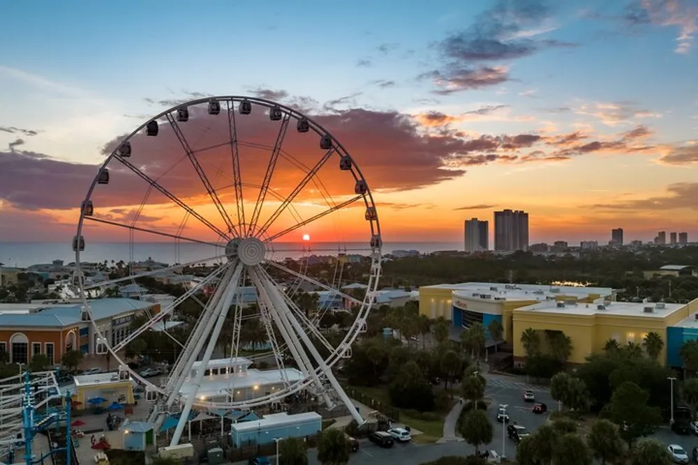 A large Ferris wheel stands against a beautiful sunset skyline with scattered clouds overlooking a coastal cityscape