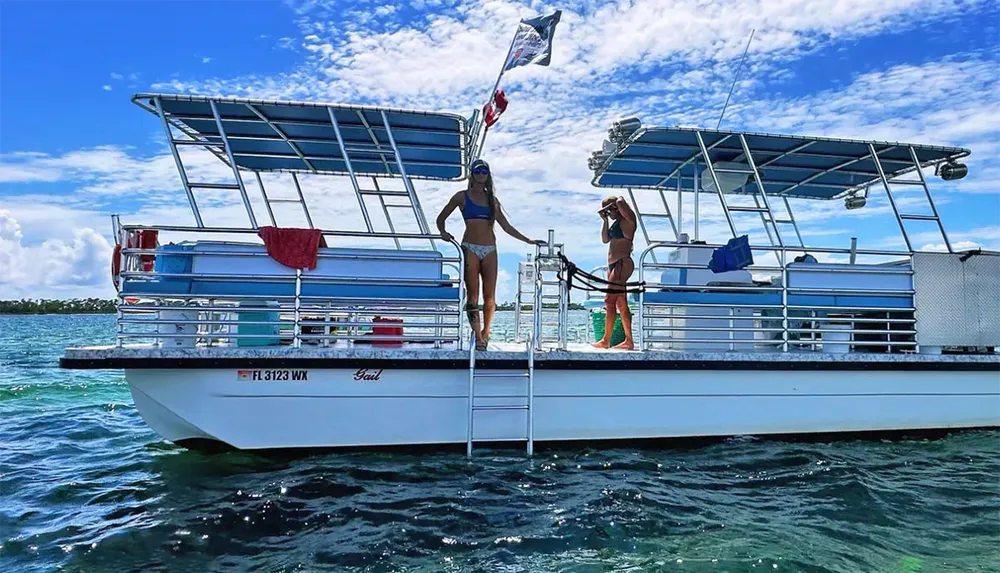 Two people are on the rear deck of a pontoon boat one about to descend a ladder into the water and the other standing and holding a beverage against a backdrop of clear skies and calm seas