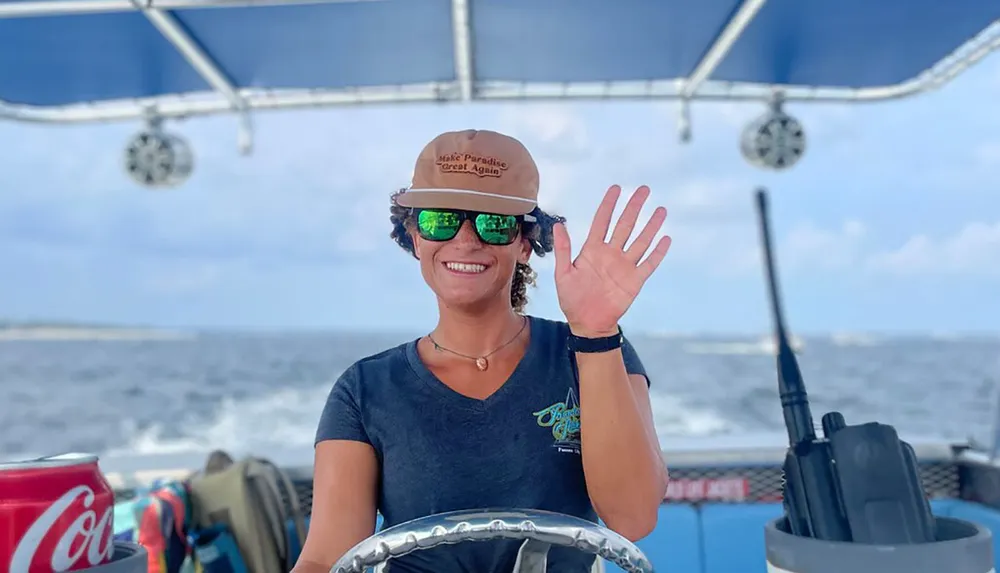 A person wearing a cap and sunglasses is smiling and waving at the camera while steering a boat