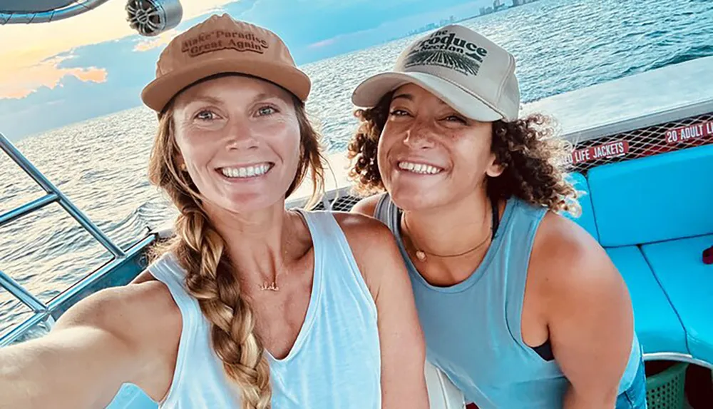 Two smiling people are taking a selfie on a boat with one person wearing a cap with the phrase Make Paradise Great Again and the other wearing a cap that says Conserve Wildlife