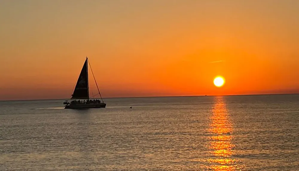 A sailboat cruises on calm waters against the vibrant backdrop of a setting sun reflecting on the sea