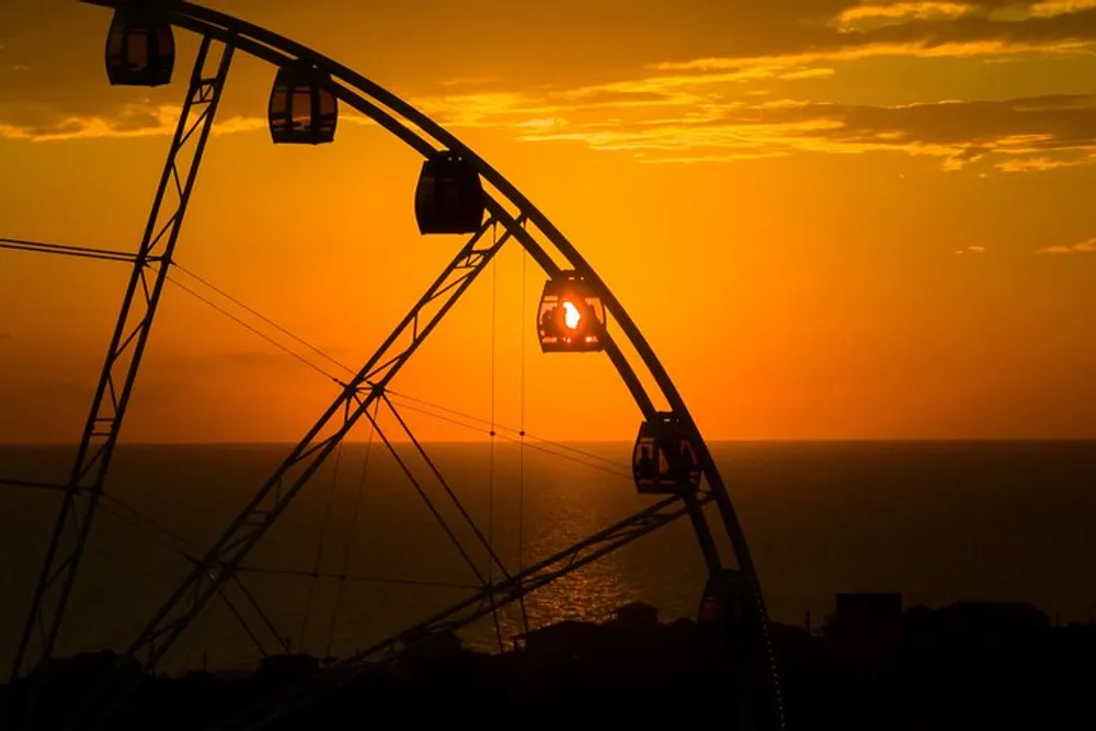 A silhouette of a Ferris wheel stands against a vibrant sunset over the ocean