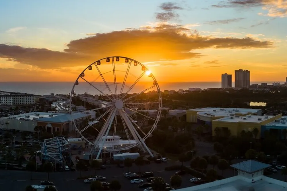 A scenic view of a Ferris wheel silhouetted against a vibrant sunset sky with the ocean in the background and a parking lot in the foreground