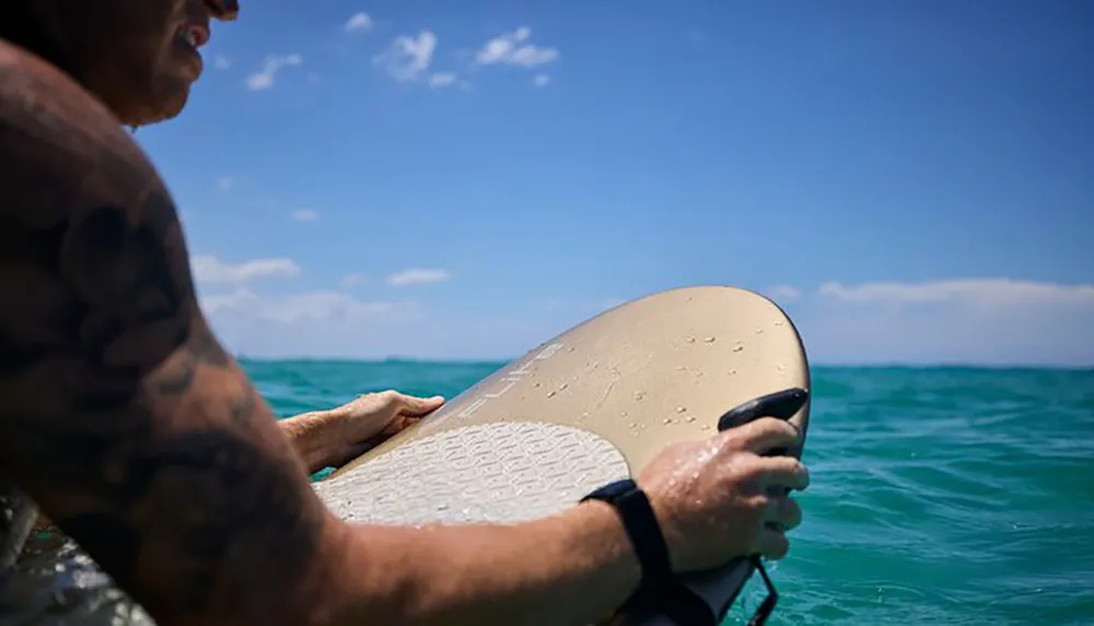 A person with a tattooed arm is seen lying on a surfboard in clear blue ocean waters holding onto the board with a leash attached to their wrist