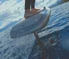 A persons feet are on a surfboard cutting through the water with mountains and a blue sky with clouds in the background