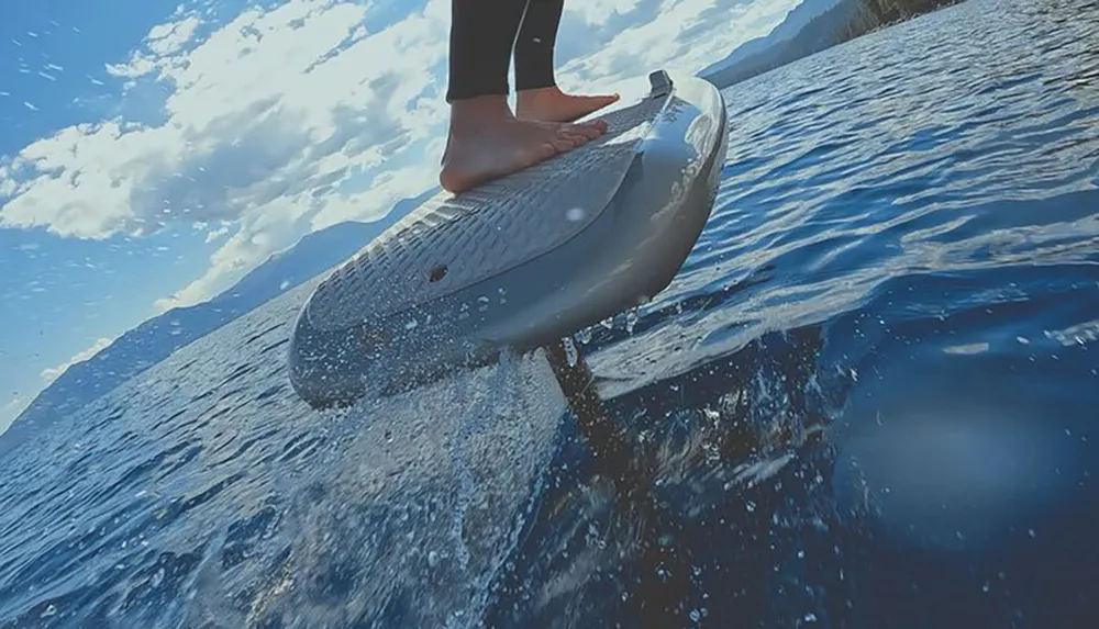 A persons feet are on a surfboard cutting through the water with mountains and a blue sky with clouds in the background