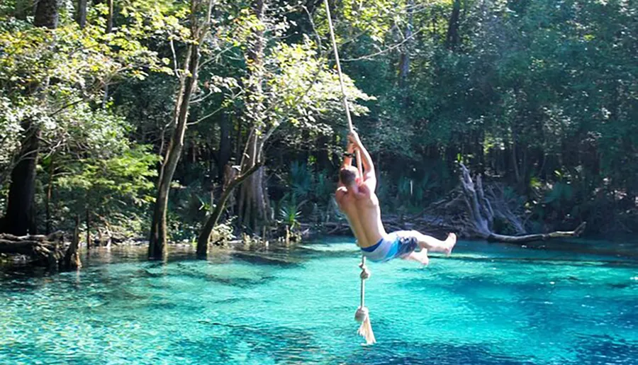 A person is joyfully swinging on a rope above a clear blue water body surrounded by lush greenery.