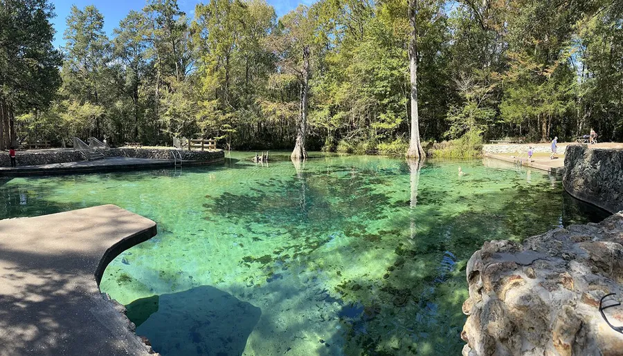 The image shows a serene, crystal-clear natural spring surrounded by lush greenery, with people enjoying the water and the natural setting.