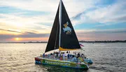 A sailboat with a black sail bearing a pelican image is cruising on the water at sunset with a group of passengers enjoying the scenery.