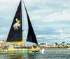 A colorful catamaran with a pelican graphic on its sail is gliding through the water near a coastal area with buildings in the background