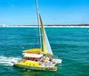 A yellow catamaran with a group of people on board is sailing on the blue ocean near a coastline with distant buildings under a clear sky