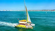A yellow catamaran with a group of people on board is sailing on the blue ocean near a coastline with distant buildings under a clear sky.