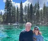 A smiling couple poses for a photo with a backdrop of crystal-clear turquoise water and a lush pine forest under a blue sky
