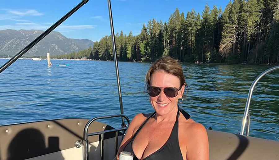A woman in sunglasses is smiling on a boat with a backdrop of clear water and forested mountains.