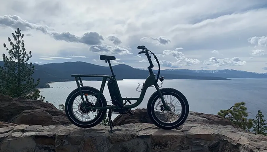 A bicycle stands silhouetted against an expansive view of a lake and mountains under a partly cloudy sky.