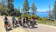 A group of people are posing with their bicycles on a sunny day near a picturesque lake surrounded by mountains and trees.
