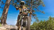 A dirt bike rider in full gear takes to a dusty trail amidst a wooded area under a clear blue sky.