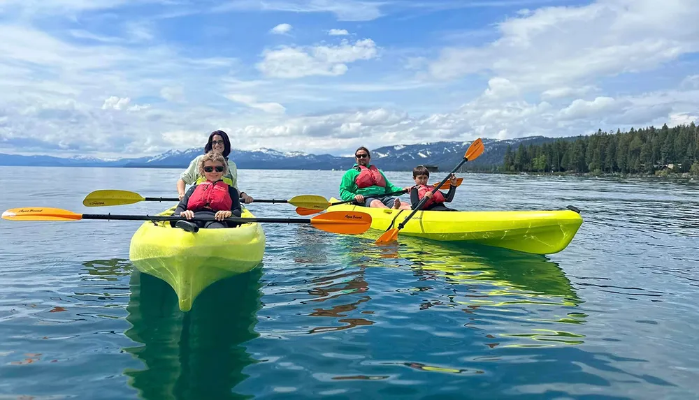 Four people are kayaking on a calm lake with mountains in the distance