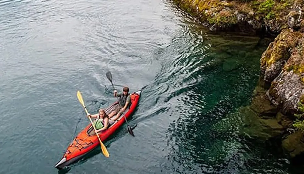 Two people are kayaking in clear tranquil waters near a rocky shoreline