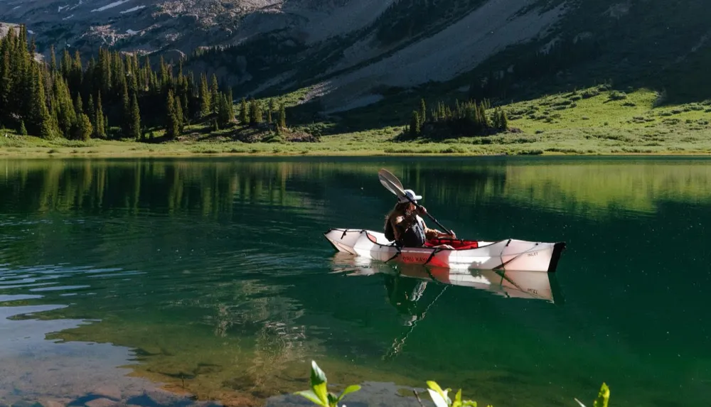 A person paddles a kayak on a tranquil mountain lake surrounded by forested slopes