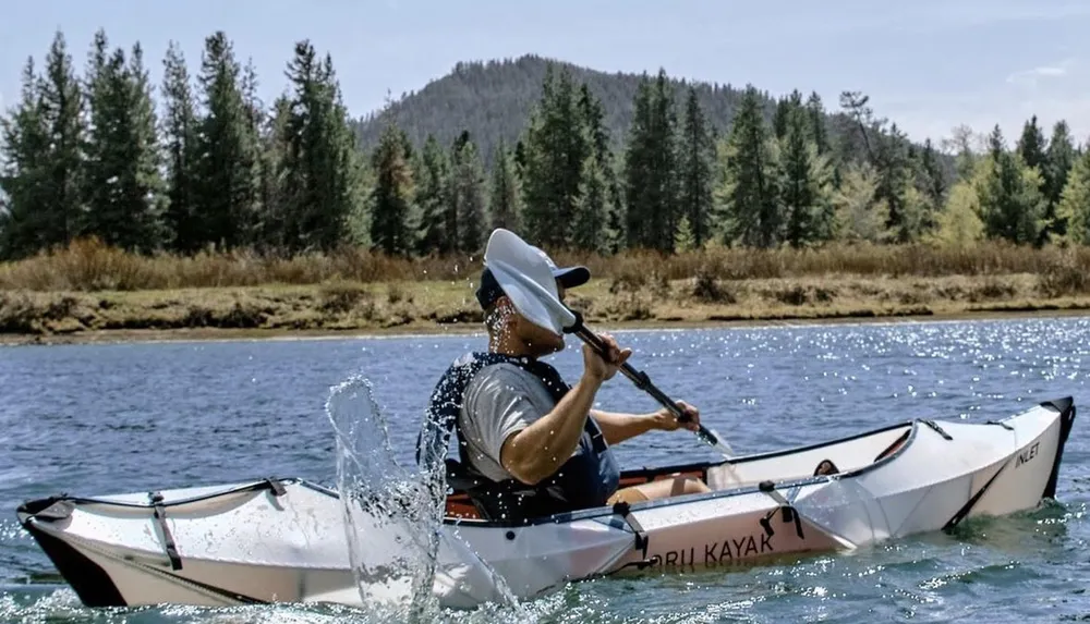 A person in a white sun hat is kayaking on a shimmering body of water surrounded by a forested area