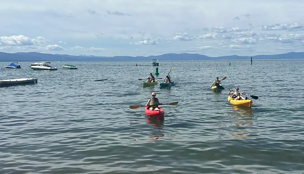 A group of people are kayaking on a calm lake with anchored boats and a mountainous horizon in the background