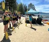 Two smiling people one holding a paddle and the other holding a dog are seated in a bright yellow kayak near a beach with other colorful kayaks and beachgoers in the background