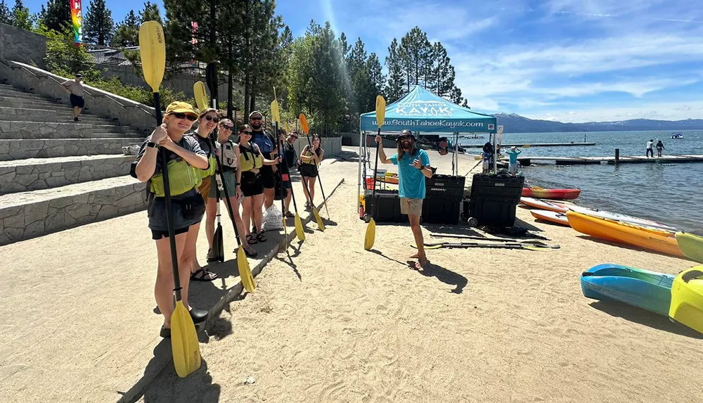 A group of people holding kayak paddles stands on a sunny beach with kayaks nearby ready for a water adventure