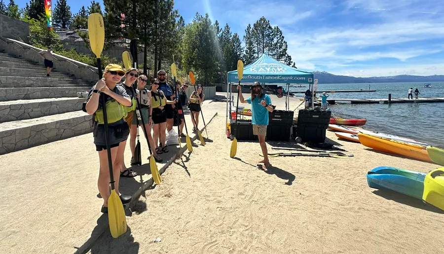 A group of people holding kayak paddles stands on a sunny beach with kayaks nearby, ready for a water adventure.