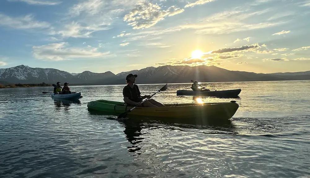 Three people are kayaking on a tranquil lake against a backdrop of mountains and a setting sun