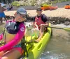 Two smiling people one holding a paddle and the other holding a dog are seated in a bright yellow kayak near a beach with other colorful kayaks and beachgoers in the background