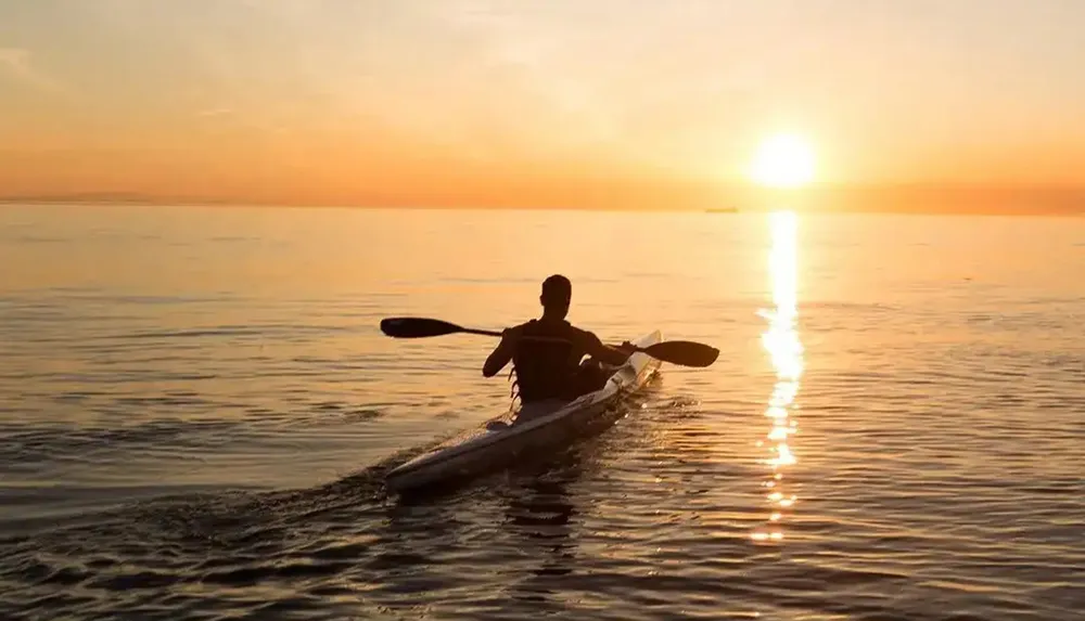 A person is kayaking on calm water during a beautiful sunset creating a path of light on the waters surface