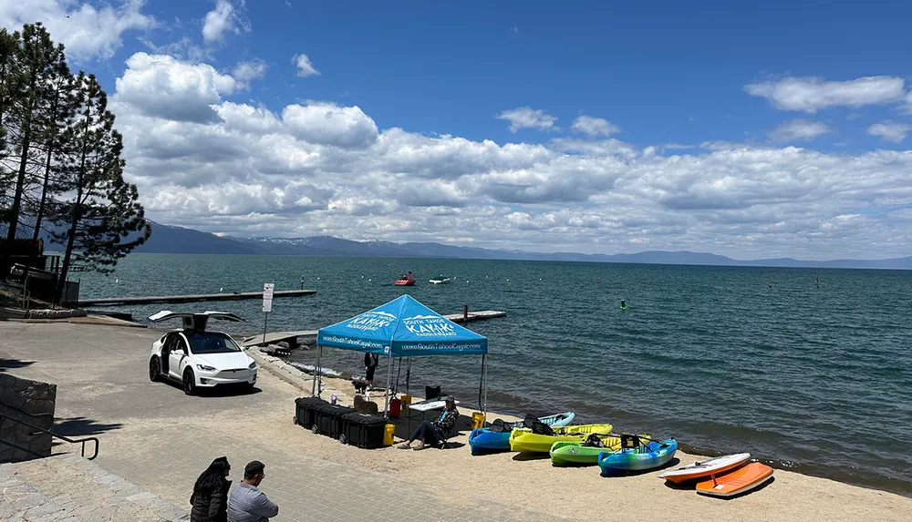 The image shows a scenic lake view with colorful kayaks on the shore a watersport rental tent and some people enjoying the waterfront with mountains in the background on a partly cloudy day