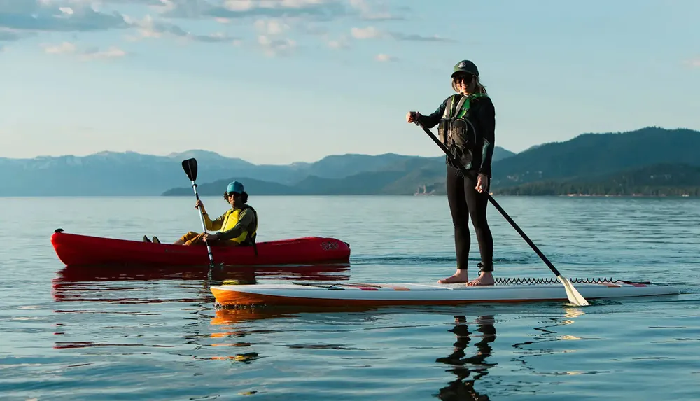 Two people equipped with life jackets are enjoying water sports with one sitting in a red kayak and the other stand-up paddleboarding on a calm lake with mountains in the background