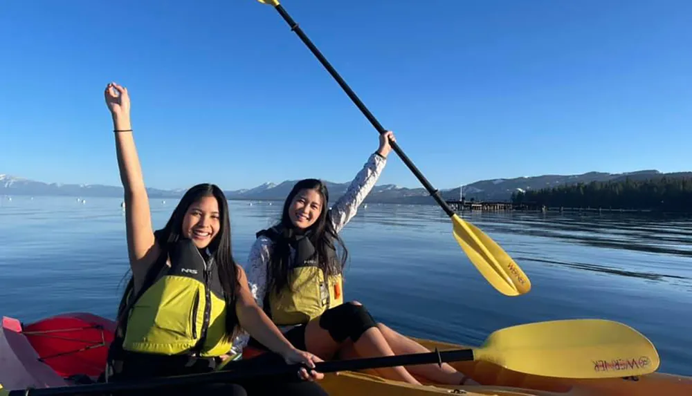 Two people are smiling and posing with a kayak paddle on a serene lake with mountains in the distance