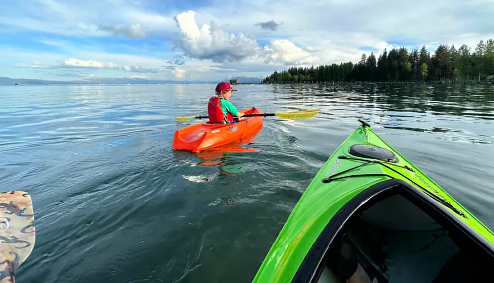 A person in a red kayak paddles on a calm lake with another green kayak visible in the foreground